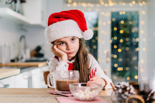 Bored cute little girl kid in Santa Claus hat sitting on the decorated for winter holidays modern kitchen with cocoa drink. Festive season. Copy space.