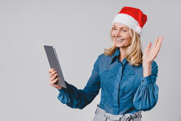 Videocall conference meeting. Mature middle-aged businesswoman in Santa Claus red hat using digital tablet for online shopping for Christmas New Year, texting, surfing social media on grey background