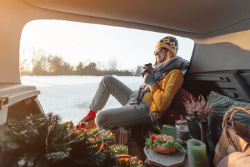 Woman with coffee in hands is sitting in car trunk and has winter picnic