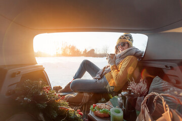 Woman with coffee in hands is sitting in car trunk and has winter picnic © Oleksandr Kozak