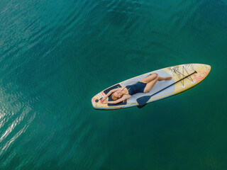 Fototapeta premium A woman lies on a sup, view from a drone. Young women Having Fun Stand Up Paddling in the sea. SUP. Red hair girl Training on Paddle Board near the rocks