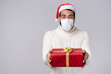 Arabian Middle-Eastern young man in protective face mask against Coronavirus Covid19 and Santa Claus red hat giving Christmas New Year present gift isolated over grey background