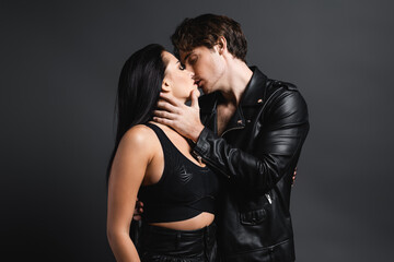 man in leather jacket kissing sexy woman in black crop top on grey.