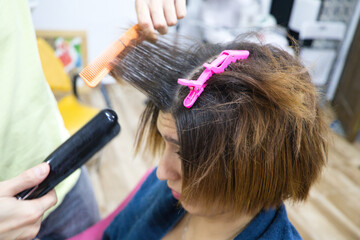Fototapeta premium Transgender woman combing her hair with hairdryer, comb and hair straightener in the beauty salon by the professional stylist. Concept of beauty and hairdressing. Beauty and health center.