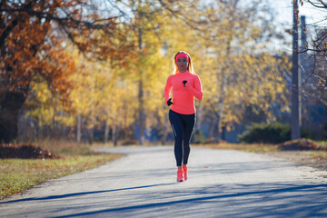 Young fitness woman running on the road in the cold morning in winter. Sporty girl in orange long-sleeve jogging in the sunny morning