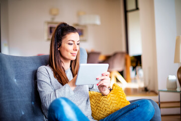 Happy attractive woman using her digital tablet while relaxing on the sofa at home