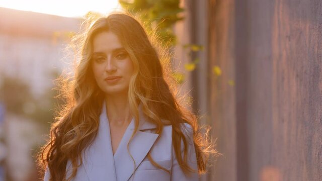 Close-up female model pensive beautiful girl with long curly blonde hair stands outside sunbeams sunset background looking at camera. Young dreamy woman posing outdoors in sunlight in windy weather