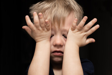 Fototapeta premium A 3-year-old blond boy cries and wipes his eyes and tears with his hands, an offended child
