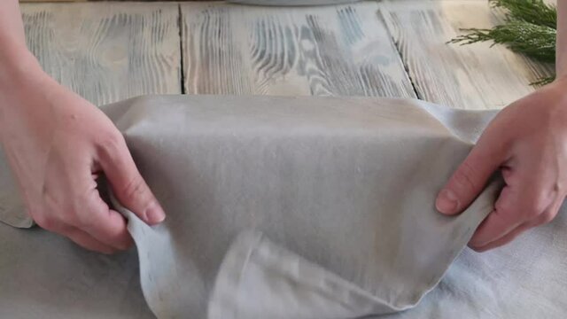Eco-friendly and zero waste concept, female hands wrapping a Christmas present in a reusable gray cloth on a white wooden background, furoshiki.