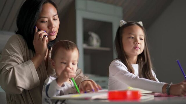 Smiling Asian mom talking by phone and looking after children in the kitchen