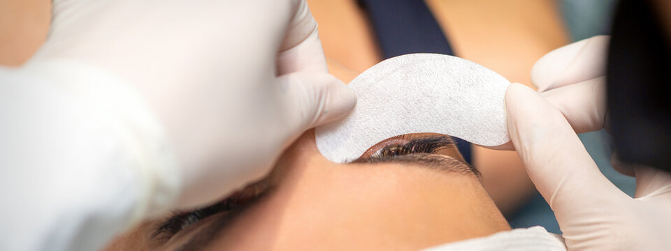 The beautician is gluing a patch under her eyes, before the procedure of eyelash extension. Artificial extended eyelashes