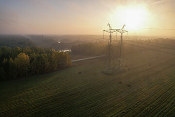 AERIAL VIEW, LENS FLARE: Support of a high-voltage power transmission line with wires in a wooded rural area opposite the rising sun. Transmission of electricity over long distances.