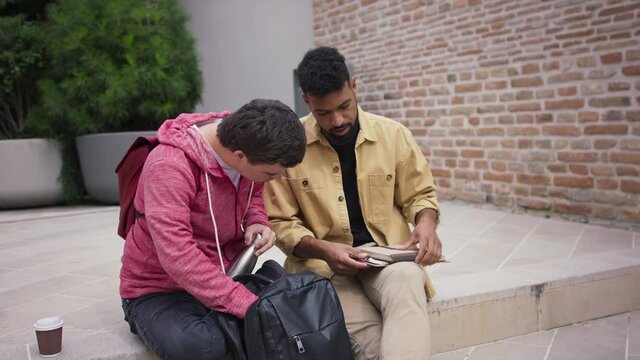 Happy young man with Down syndrome and mentoring friend sitting and talking on stairs outdoors in campus area.
