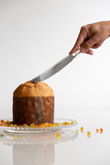 Panettone being sliced, next to colorful candied fruits, in a photo on a white glass table and...