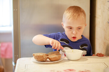 A little boy with a genetic disease Down Syndrome is sitting at the table and eating. The child...