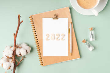 Top view Desktop Christmas notepad with 2022 text. Flat lay on green mint table background with planner, cup of coffee, tree brunch, Christmas eco nature decoration, notebook and stationery.
