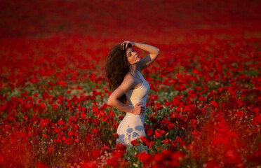 Beautiful slim, long haired brunette woman in white dress stands in the middle of a red flower field. 