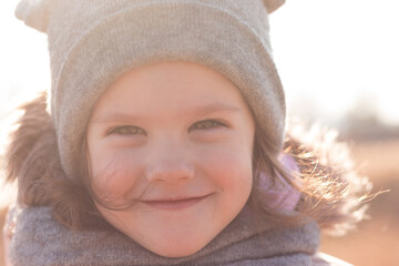 Close-up portrait of a smiling little child in a warm hat with ears in the sunlight on a walk in the park