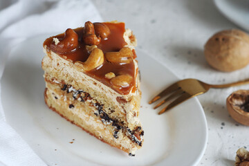 A piece of nut cake is in a plate on a light table. Nearby lie a golden fork, nuts and a towel