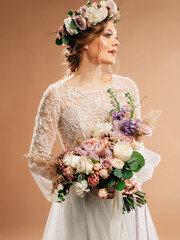 Gorgeous happy bride in white wedding dress in brown studio with floral wreath and large purple bouquet. Smiling blonde lady in white transparent lace gown, bridal fashion. Wedding floristic trends.