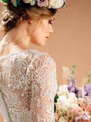 Beautiful blonde bride with stylish hairstyle in a floral wreath with bridal bouquet,back view. Bridal portrait in studio with brown background. Wedding fashion, white transparent lace gown close up.