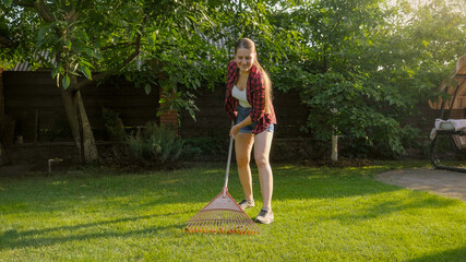 Young smiling woman working at house backyard and collecting fallen leaves with garden rake....