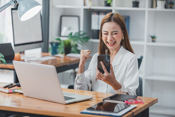 Excited Asian woman sitting at her desk Feel satisfied with the business going well. Very happy to receive letters from laptops and smartphones promoted at work.