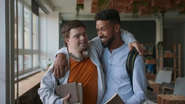 Young man with Down syndrome and his tutor hugging and looking at camera indoors at school.