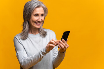 Naklejka premium Smiling senior woman holding and using smartphone, carefree mature modern gray-haired lady enjoying online chatting in social media, using mobile app, isolated over yellow background
