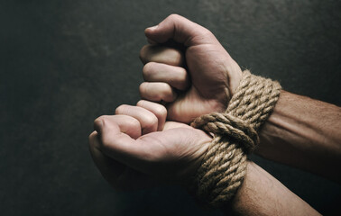 Male hands tied with a rough rope at the wrists close-up on a dark background, soft focus....