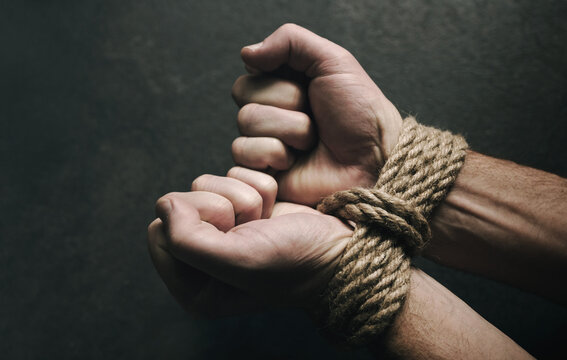 Male hands tied with a rough rope at the wrists close-up on a dark background, soft focus. Conceptual image of violence, slavery, dependence, lawlessness, lack of freedom