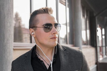 Portrait of a serious young guy of European appearance, an attractive businessman in sunglasses, listening to music in headphones on the street outdoor