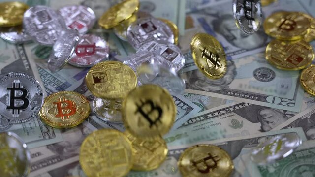 A slow motion view of dropping golden and silver Bitcoins over rotating US currency.	