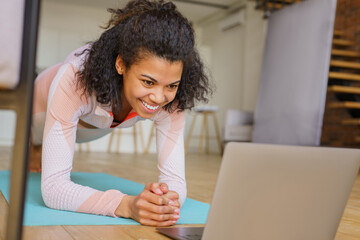 Beautiful smiling fitness woman using laptop watching online tutorial, doing exercises standing in plank pose at home. Sport, healthy lifestyle concept  