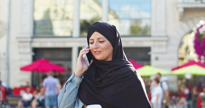 Adult female arab wearing hijab, walking on street, talking on cellphone. Muslim woman with veiled head calling friend, holding coffee cup, smiling. Communication, technology, religion concept.