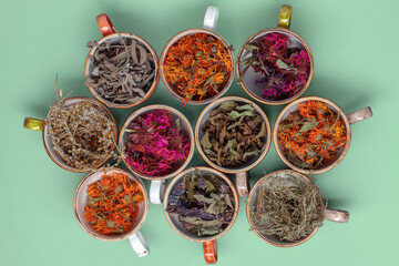 Fototapeta premium Assortment of dried relaxing tea herbs in colourful cups on mint green background. Calendula, mint, anise hyssop, monarda didyma, wormwood, sage leaves.