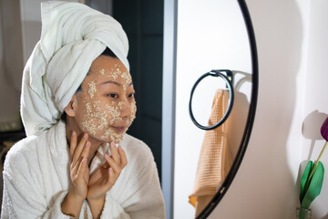 Portrait of relaxed young asian woman making oatmeal mask in the bathroom in front of the mirror.
