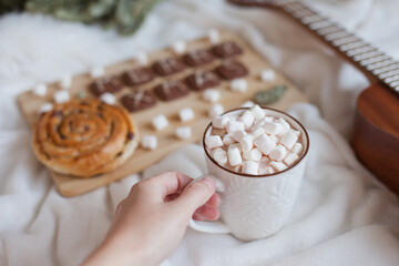 Hand holding a cup of hot cocoa with marshmallows with christmas decor and cookies. Merry Christmas cookies with ukulele and marshmallows and xmas wreath with cozy lights. Christmas at home background