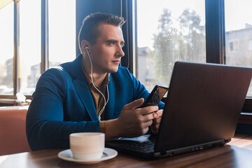 A business pensive man stylish businessman in an attractive European-looking suit works in a laptop, listens to music with headphones and sitting at a table in a cafe by the window