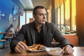 A serious, brooding stylish young man of European appearance, a businessman in a jacket sits at a table in a restaurant on a lunch break and looks out the window