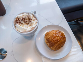 Top down view of a coffee topped with whipped cream and a pain au chocolat on a white, marble table.