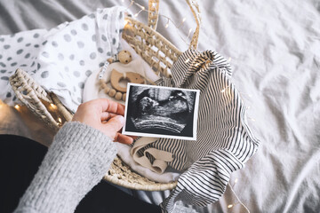 Pregnant woman with wicker basket of newborn clothes, ultrasound image, toy. Expectant mother...