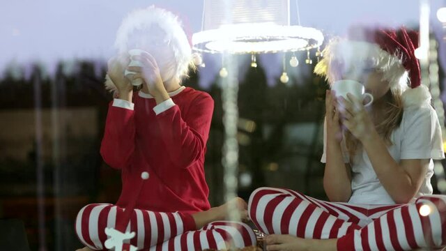Brother and the sister dressed in Santa’s hat are having their tea break in Christmas decorated environment. Filmed through the glass window
