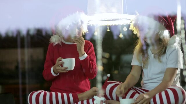Brother and the sister dressed in Santa’s hat are having their tea break in Christmas decorated environment. Filmed through the glass window