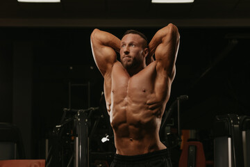 A bodybuilder with a beard is doing a stomach vacuum pose during a workout in a gym.