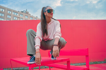 Cool stylish young girl listen to rap music with wireless earphones squat on table outdoors in...