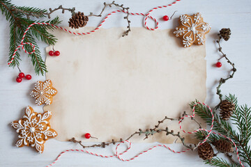 Christmas white wooden background with old paper for congratulation text, list, letter to Santa and fir branches, gingerbread cookies snowflakes, cones, red berries, red and white rope.