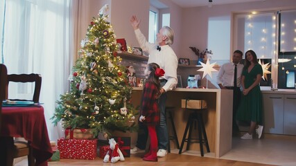 Happy grandfather and granddaughter hanging decorations at Christmas tree at Christmas or New Year party at home. Xmas celebration preparation or winter holidays.