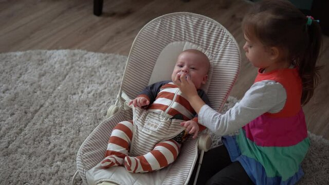 Childhood, motherhood, family, comfort. Little newborn baby boy in striped bodysuit pajamas awake looking around smiles plays rattles sibling sister kid lul amuse brother in room at home atmosphere