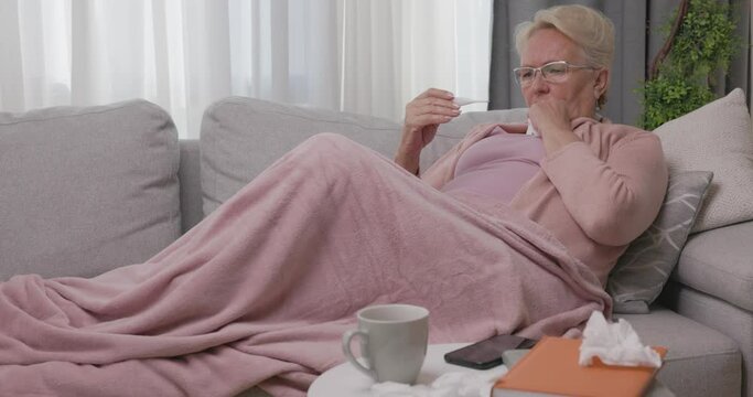 Sick elderly woman checking her temperature with thermometer lying on sofa caughing suffering from seasonal flu or cold. Ill mature senior feel unhealthy with influenza at home